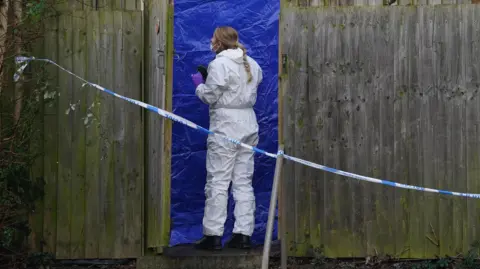 PA Media A forensic officer wearing white overalls standing in front of a blue tent with green fencing either side and blue and white police tape across the whole scene