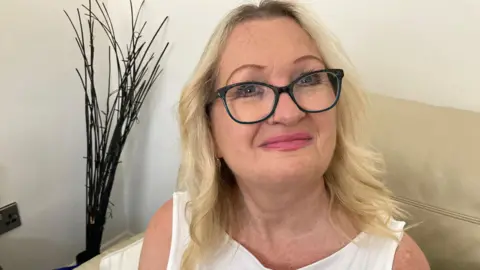 A woman with blonde hair smiles at the camera. She is wearing glasses and a white top. She is sat on a cream sofa. 