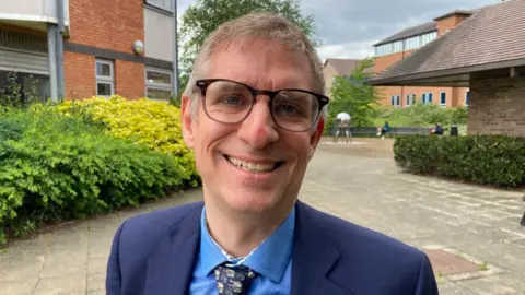 Councillor Matt Jenkins, wearing a blue suit jacket, blue shirt and patterned tie, is smiling at the camera.