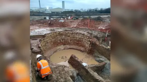 Cotswold Archaeology The remains of a Roman limekiln - a curved wall with muddy water in it - at an archaeological dig site. An archaeologist wearing an orange hi-vis jacket and a white helmet is working near it. In the background is an industrial site with white warehouses and chimneys.
