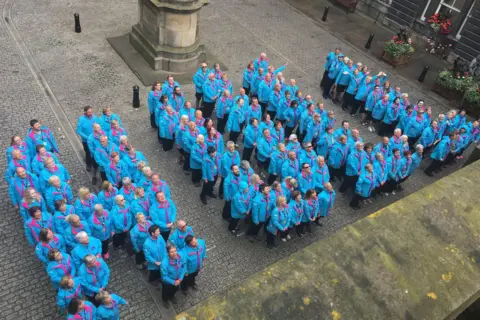 Dale Baxter/BBC A photo taken from above showing dozens of people wearing bright blue jackets and standing in a pattern that spells out the word "Hull" in a cobbled courtyard with a statue behind them.