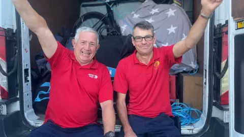 Alan and Toby wearing red tops sat in the door of a van together. They both have one arm in the air and are smiling at the camera. 