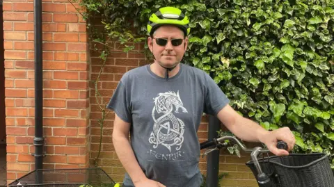 James Treacy James Treacy standing next to a bike. He is wearing a high viz helmet, sunglasses and a grey graphic t-shirt.