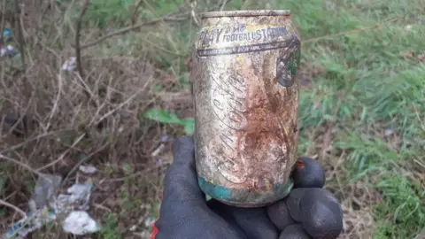 MARK FISHPOOL A faded can of Coca-Cola covered in a light layer of dirt and rust is held by a thick black glove above grass and scrubland where more litter can be seen on the ground. 