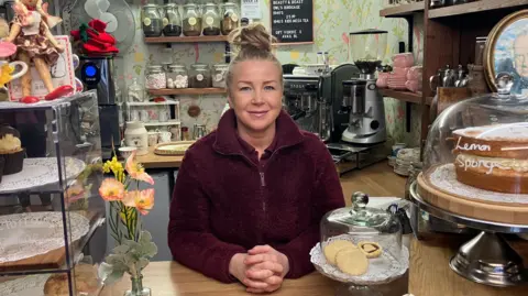 Claire Church smiling behind a cafe counter which has a vintage tea party theme. She is framed by sponge cakes and biscuits with shelves full of loose leaf tea behind her. She has blonde hair tied in a high bun and wears a maroon fleece jacket over a similar coloured collared shirt.