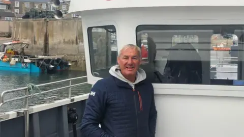 Andrew Douglas is standing by his boat in Seahouses harbour. He is a man in his 50s wearing a blue coat with a white hoodie and has short blondish grey hair.