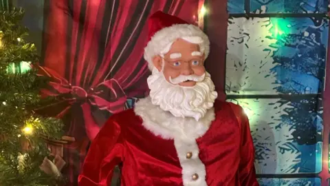 Rob Hardwick A model of a man with a beard in a red hat and coat, trimmed with fur, standing beside a Christmas tree