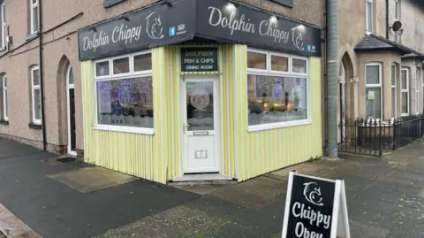 External view of Dolphin Chippy which has a black sign and yellow walls and white windows and door and sits on the corner of Blakiston Street. There is a black and white A-board sign outside saying Chippy Open.