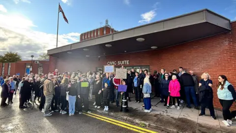 A crowd of protesters stand outside a red brick building holding up various placards. There are about 50 on the pavement and the road which has double yellow lines on it. 