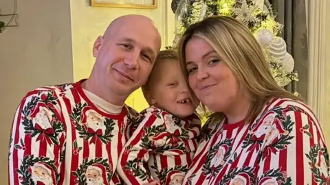 A bald man and a woman with shoulder length blonde hair are smiling at the camera, with a small child between them. All three are wearing Christmas red and white outfits