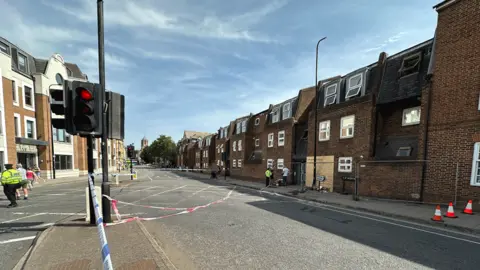 Looking down St Aldate's, a main road through Oxford. Thames Street is to the left, the damaged building opposite it. A board covers the damage. The police cordon winds around the scene.