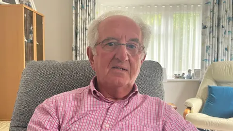 Julia Gregory / BBC A man wearing a pink checked shirt sat in a grey fabric chair.