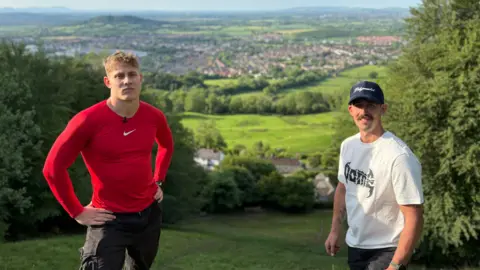 Tom Kopke, a tall blond man wearing a tight red Nike top and black shorts, stands next to a shorter man with a brown moustache, wearing a black cap, a white T-shirt, and black shorts, on the summit of a hill in rural England.