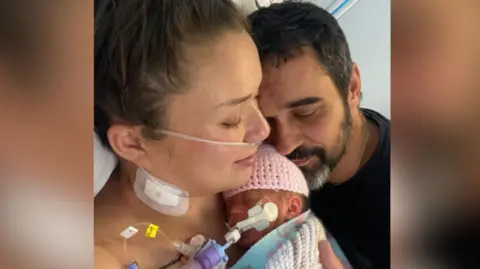 Kimberley Newark holds her baby in her arms, the baby is wrapped in a blanket and woollen hat and is connected to various medical tubes. The baby's father, Yann Trupiano is holding his face close to his daughters head with his eyes closed.