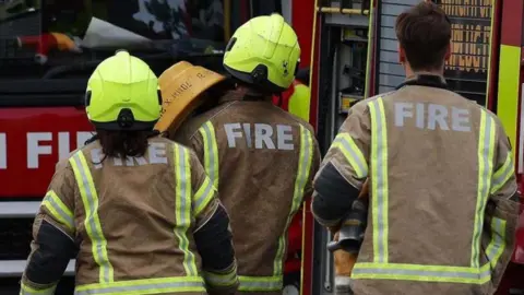 The back of 3 firefighters in uniform walking towards a fire brigade