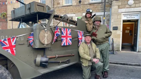 Three men in green army overalls standing next to a World War Two military vehicle. The truck is painted in khaki green and has Union Flags hanging along it.  