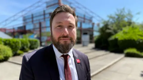 Steve Hubbard/BBC A picture of Milton Keynes Council leader Pete Marland. He is standing in front of The Point building on a blue sky day. He has a beard and short dark brown hair and is wearing a navy blue suit jacket and a wine red tie. He has a Labour pin badge on his lapel.