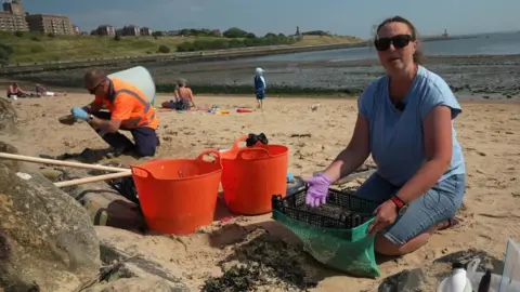 Elaine Wilson, a woman wearing sunglasses, is holding a basket, which has a green net attached to the bottom, where the nurdles and sand are collected. Next to her are two orange buckets, and further back, another man in an orange high-vis jacket helps with the clean-up.
