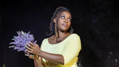 Mark Senior Georgina Onuorah as Fiona MacLaren in Brigadoon at Regent's Park Open Air theatre in London. She is looking to the right of the camera and is wearing a light yellow dress. She is holding sprigs of lavender in her hands.