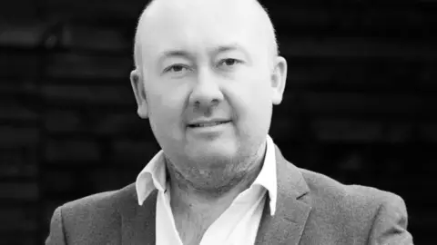 Targa Florio Cars A black and white photo of William Kirkham. He is looking at the camera and wearing a white shirt with a suit jacket.