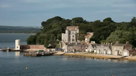 A wide shot of Brownsea Island. Several houses can be seen on the island and Brownsea Castle looms over them from behind.