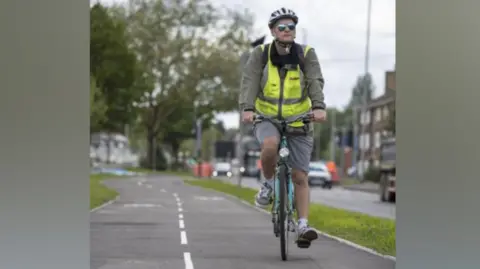 City of Wolverhampton Council A cyclist wearing a helmet and a hi-vis top is cycling on a track on the central reservation.