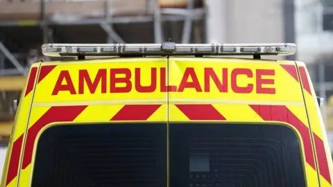 A picture of the back of an ambulance. It has yellow and red stripes. There is, in red writing, the word AMBULANCE. There is the lights attached to the top of the vehicle.