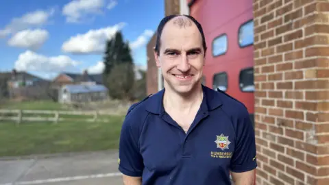 A man smiling, wearing a blue fire and rescue shirt with receding black hair stood outside a fire station near a brick wall.