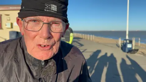 Close up image of elderly man wearing glasses and a black woolly hat on a seafront in sunshine