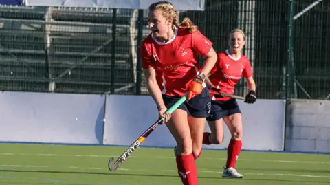 Jersey Sport Two women are playing hockey wearing red shirts, blue shorts and red socks
