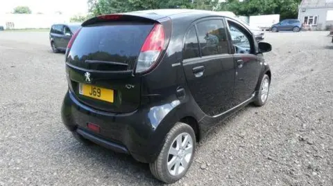 A midshot of the Peugeot Ion facing away. It is black, and has a number plate with J69 on it.