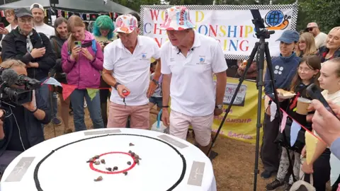 Two older men in white polo shirts and colourful bucket hats lean over a table with a white sheet, with snails on it. People with cameras are crowding around them.