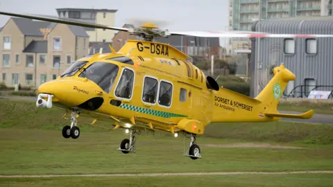 Getty Images A large yellow helicopter is pictured in flight. It is very close to the ground and either just taking off or landing. The words Dorset and Somerset Air Ambulance are printed on the vehicle's tail. A road and some buildings - presumably houses or flats - can be seen in the background, as well as a more industrial-looking building closer to the helicopter. 