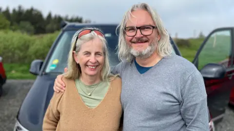Beate and Heiner Delbach are standing outside in front of a car. Heiner who has long grey hair, a beard and is wearing glasses and a long-sleeved top, has an arm around Beate. Beate also has long grey hair, has sunglasses on top of her head and is wearing a brown jumper over a green top.