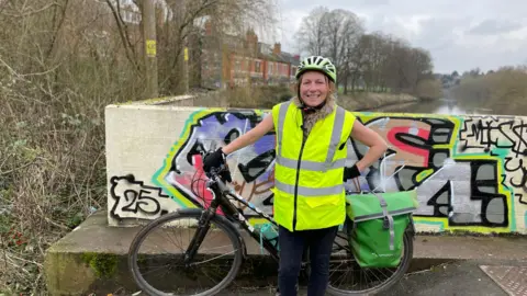 A woman wearing a yellow high-vis yellow helmet and black trousers, stands holding her bike behind her, with one hand on the handlebars and one hand on her hip. Behind her is a wall with colourful graffiti and the River Severn.