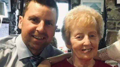 Watson family Fergal Watson pictured with his mother some years ago, before her demenita deteriorated. He has short hair and is wearing a light blue shirt and checked blue tie. She has short, blonde hair and is wearing a red jumper. 