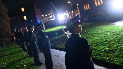 West Yorkshire Police A line of people wearing uniforms and caps standing in a row with their hands behind their backs on a pathway near a grassed area. The scene is outdoors at night.