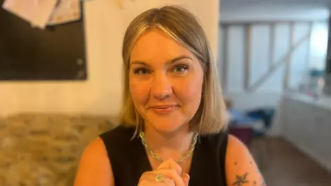 Rosie Moss is smiling and looking to camera, she's wearing a black top and gold jewellery, with short blonde hair.