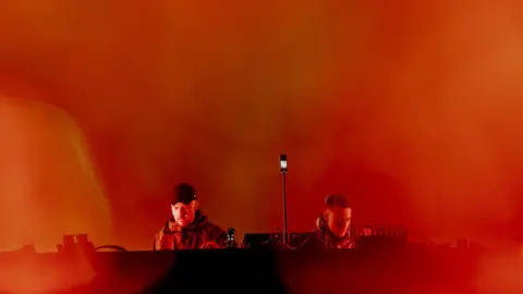 Alastair Brookes Two men, one wearing a black cap, at a black DJ deck. They are surrounded by smoky orange light.