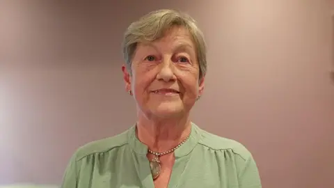 A woman, with short grey hair, wearing a green top and a necklace. 