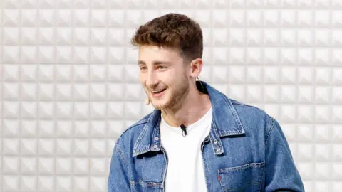 Getty Images Brandon B wears a jean jacket and white shirt. He is presenting at an event, with a microphone attached to his shirt and also on his cheek. Behind him is a simple padded white background.