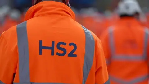 The back of a man's jacket, in hi-vis orange, with the words HS2 on them. You can't see his head. There are other people in the same jackets in front of him but they are blurry.