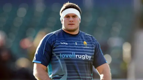 Ryan Bower wearing a Worcester Warriors shirt, standing on the pitch during a pre-match warm-up. He is wearing a white band around his head and across his ears.