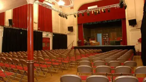 The inside of Netherton Arts Centre. The theatre has high ceilings and there are chairs set out to face the front of the small stage. 