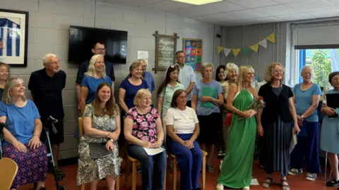 A group of men and women. Some are stood up and some are sat down. They are all smiling, inside a community centre with pictures, a TV and bunting on the wall. 