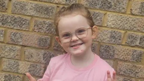 Family handout Phoebe is standing in front of a brick wall. She has ginger hair in a ponytail and is wearing a pink T-shirt. She is holding her thumbs up and smiling. 