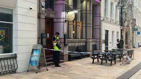 An alternative angle of the building frontage, with police tape around three black wooden benches on the pavement, and two police officers using smartphones, standing guard outside the front door. 