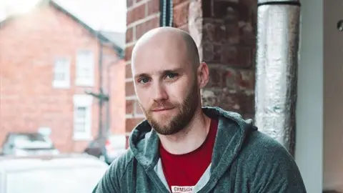 Joe Friel Shows a bald man with a dark beard wearing a red T-shirt and green hoodie with a house and cars in the background. 