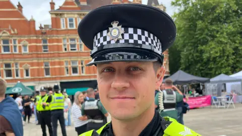 Chris Arnold  wearing a police hat stares at the camera. He is stood in Peterborough city centre square and other officers and general public can be seen behind him. The square has small marquees on it and chairs and trees in the background.