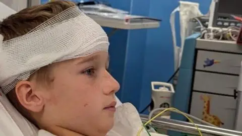 Kieran Kai, an 11-year-old boy, lying on a hospital bed with a bandage around his head